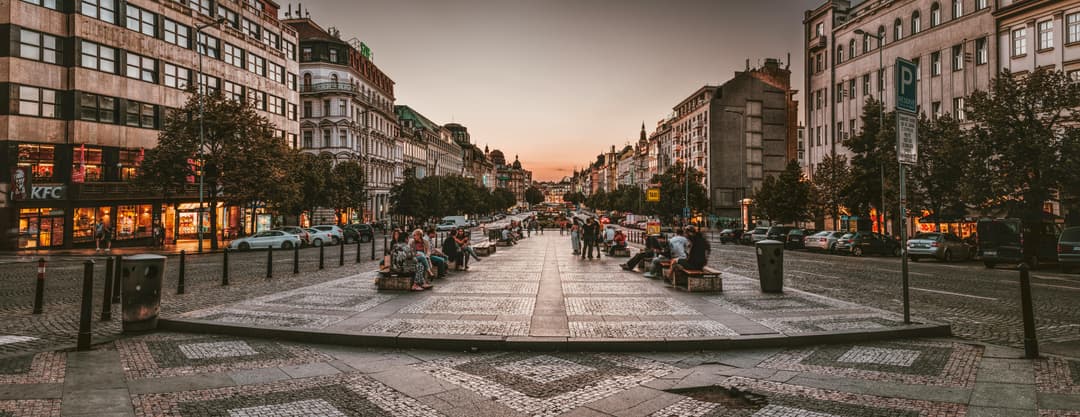 Prowall Free Wallpaper | A bustling urban street in Prague at sunset, featuring people, cars, and vibrant architecture.