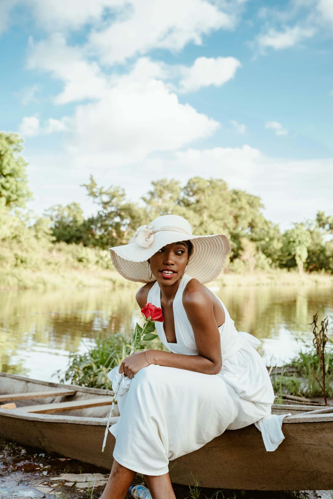 Prowall Free Wallpaper | Beautiful African woman in white dress and sunhat holding red rose, seated by a serene lake.