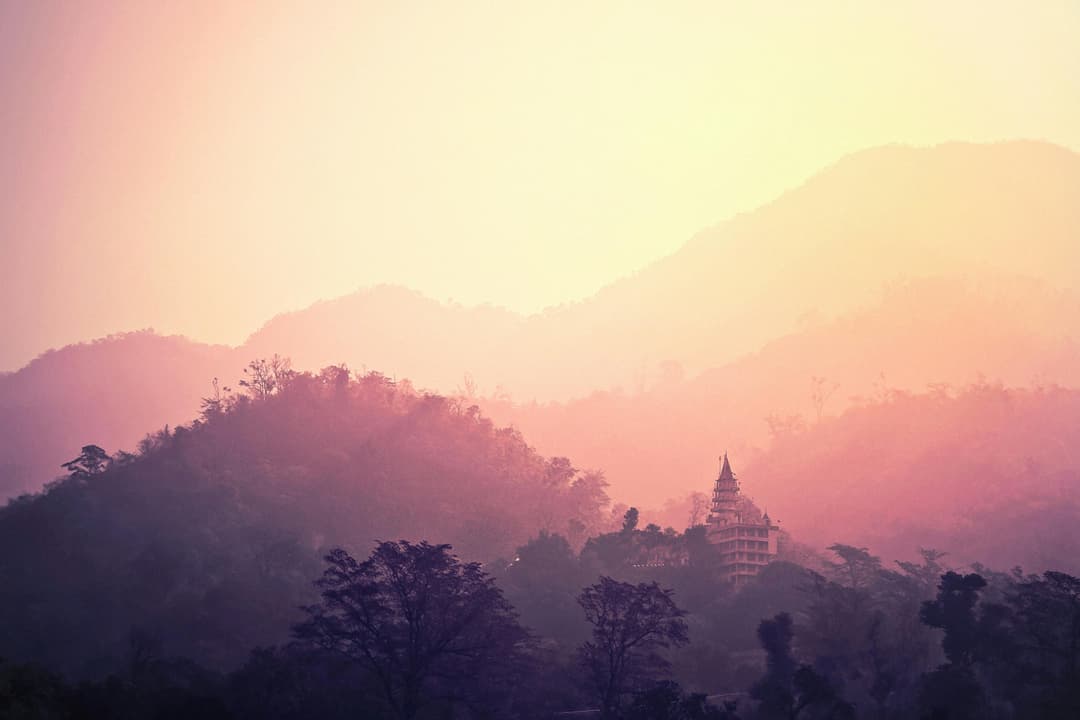 Prowall Free Wallpaper | A tranquil view of Rishikesh mountains and temple enveloped in pink hues at sunrise.