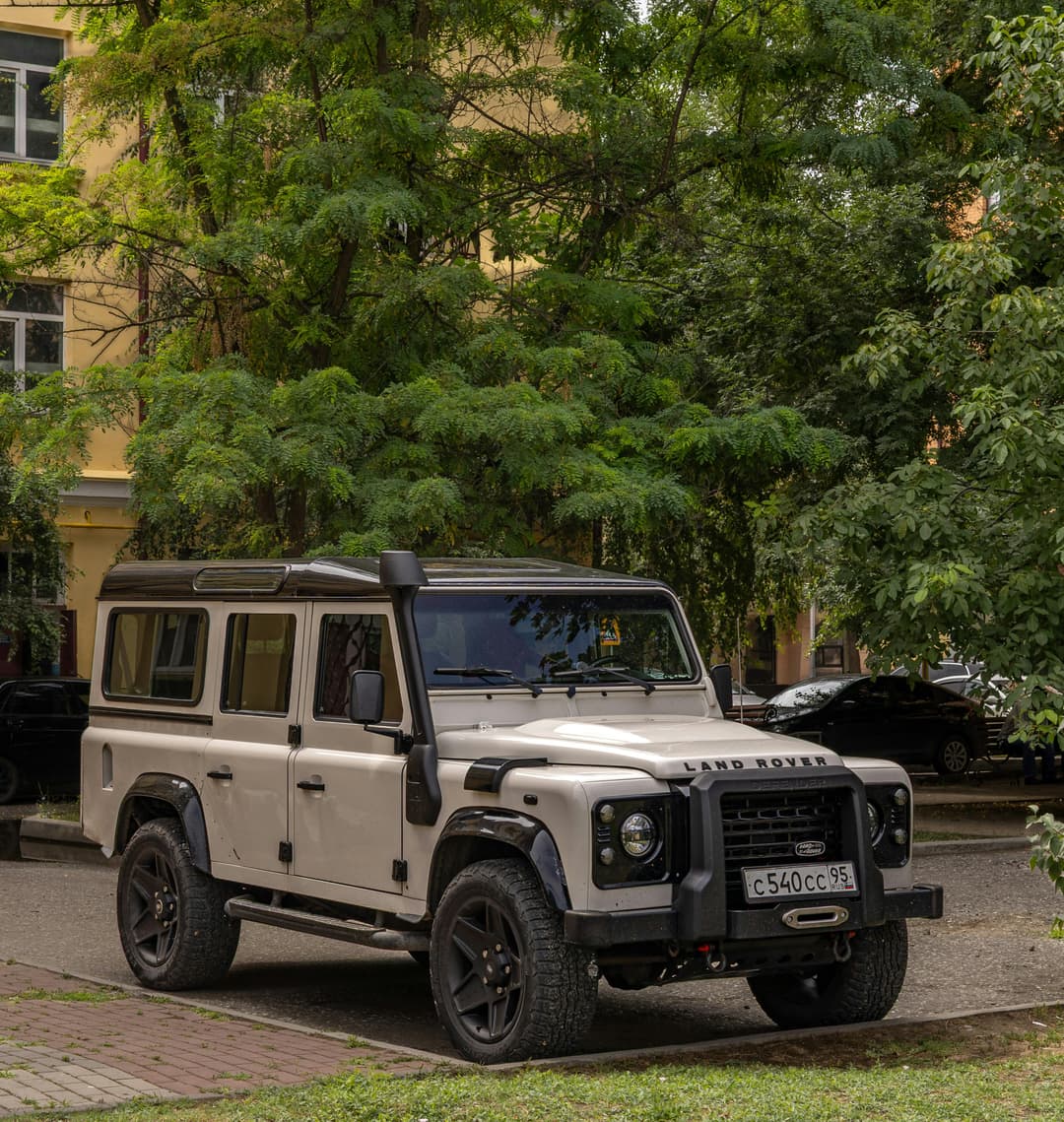 Prowall Free Wallpaper | Vintage Land Rover Defender parked under green trees on a sunny day in the city.